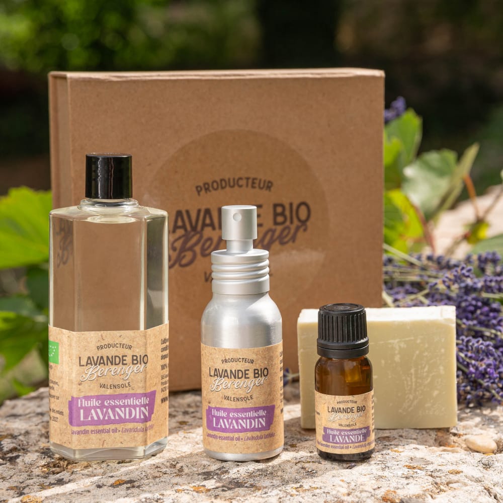 Un ensemble de produits à base de lavande biologique, comprenant de l'huile en bouteille, un spray, une petite fiole et un pain de savon, est présenté à l'extérieur avec des brins de lavande et un coffret cadeau marron en arrière-plan.