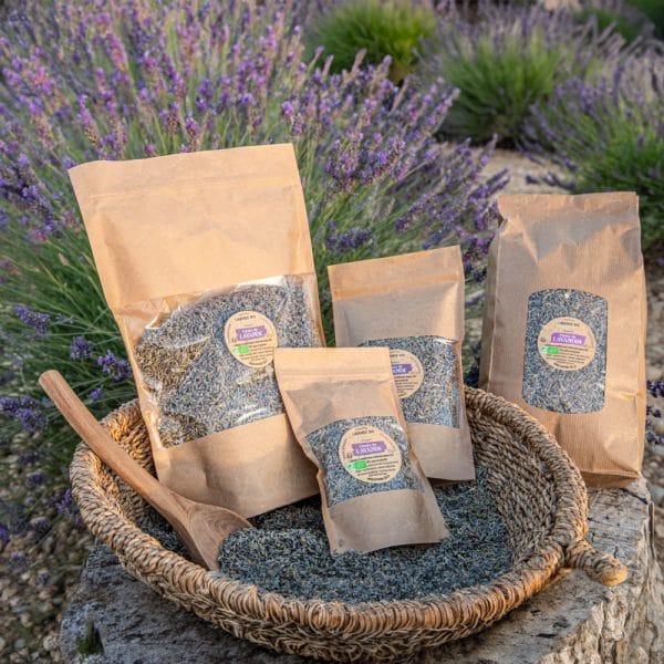 Unser Sortiment 6 Un panier tressé rempli de sachets de lavande séchée et d'une cuillère en bois est posé à l'extérieur devant des plants de lavande en fleurs. Les sacs sont de tailles différentes et portent des étiquettes.
