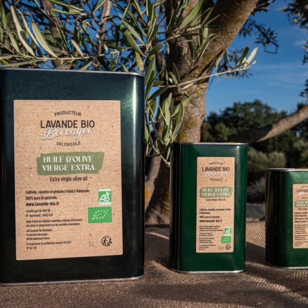 Unser Sortiment 10 Trois boîtes métalliques d'huile d'olive extra vierge biologique avec des étiquettes brunes rustiques sont présentées en plein air sur une table, avec un olivier et la lumière du soleil en arrière-plan. Les boîtes sont de tailles différentes.