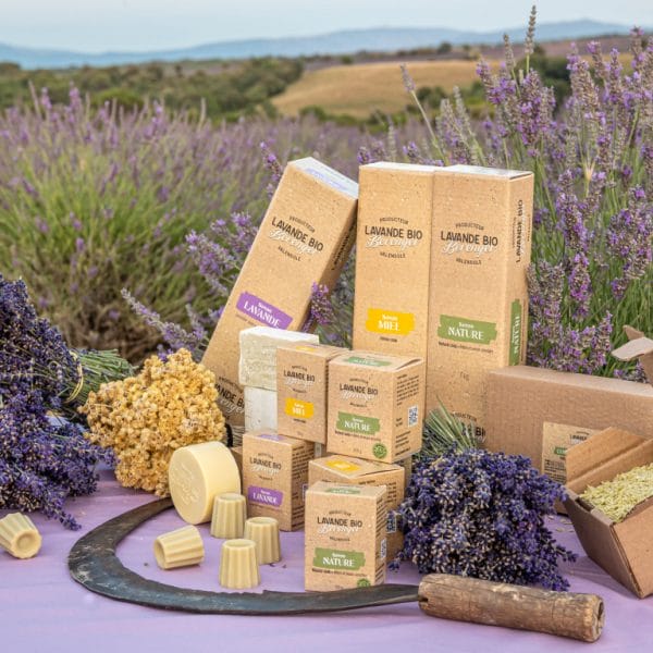 Unser Sortiment 3 Boîtes de produits biologiques à base de lavande disposées en plein air sur une table, entourées de fleurs de lavande, d'herbes et de petites barres de savon, avec une faux et un champ de lavande pittoresque en arrière-plan.