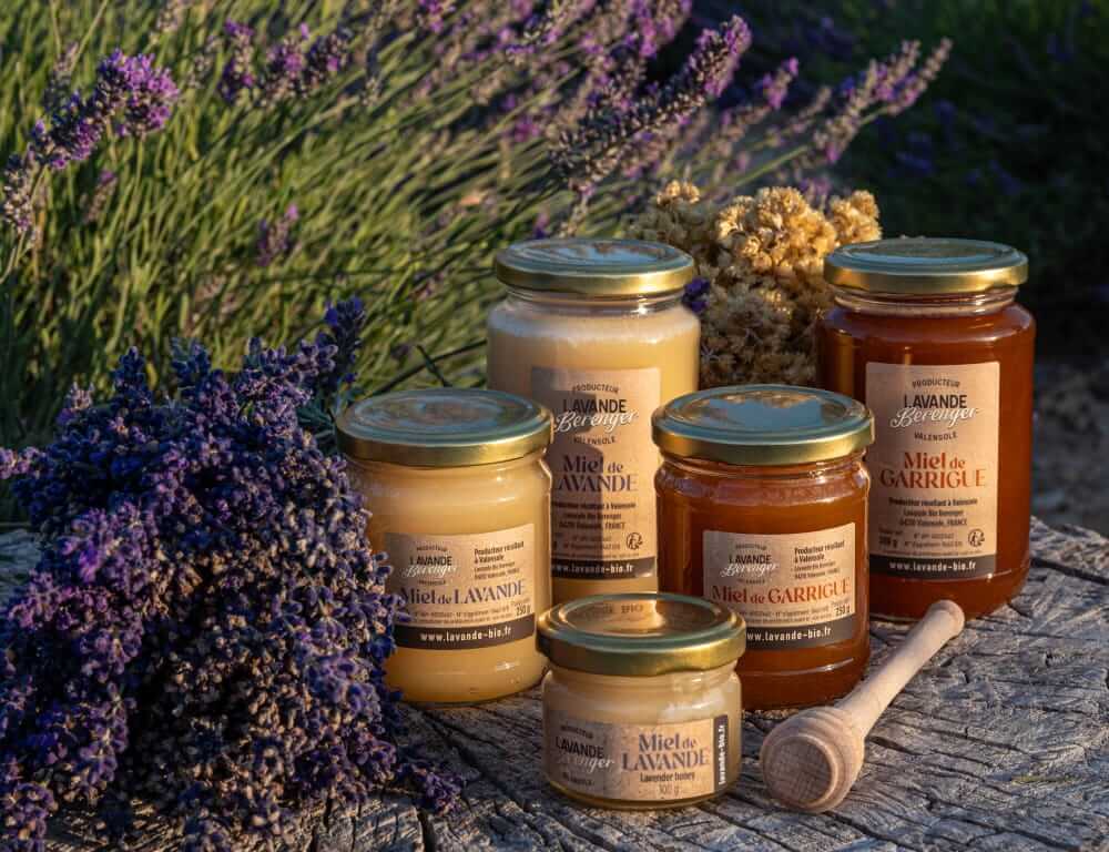 Des pots de miel étiquetés "Miel de Lavande" et "Miel de Garrigue" sont disposés sur une table à l'extérieur, entourés de brins de lavande et d'autres fleurs séchées, avec une louche à miel en bois placée à proximité.