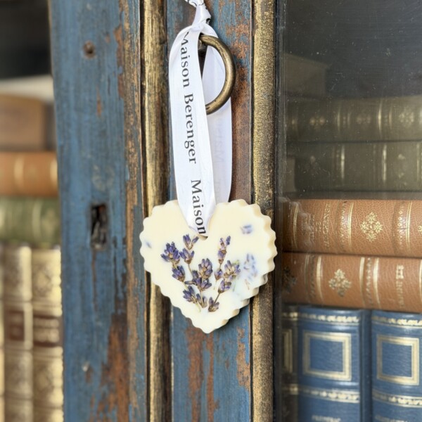 A Lavander macerate (Copie) heart-shaped ornament with dried lavender and a white ribbon hangs from a worn blue cabinet’s glass-paneled door, beside shelves of vintage books.