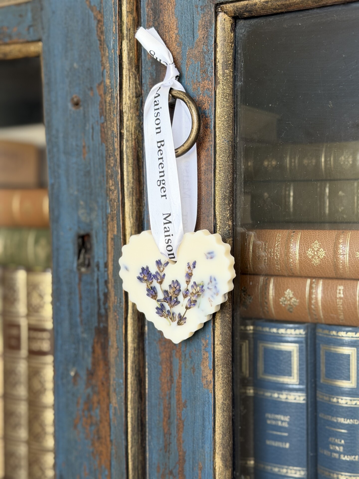 Auswahl Lavandin-Klassiker 2 Le Galet Cœur Lavande | Édition Artisanale - un ornement en forme de cœur avec des fleurs séchées au macérat et un ruban - est suspendu à un anneau métallique sur la porte d'une armoire bleue rustique, à côté d'étagères de livres anciens.